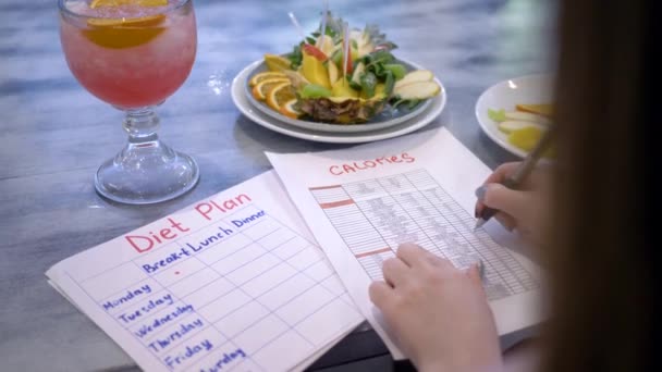 nutritionist woman writes diet plan with food calorie sheet on table with punch and fruits in plate at cafe - Footage, Video