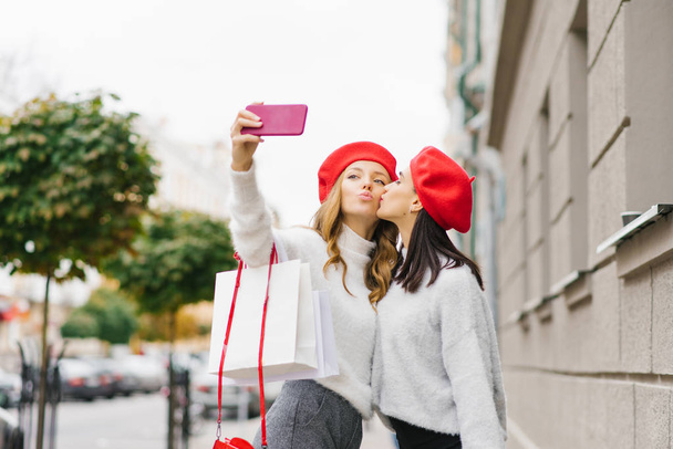 Kırmızı bereli ve açık renk kazaklı güzel kız arkadaş telefonda selfie çekiyor, kız arkadaş diğer kız arkadaşını yanağından öpüyor, kız alışveriş torbaları tutuyor. - Fotoğraf, Görsel