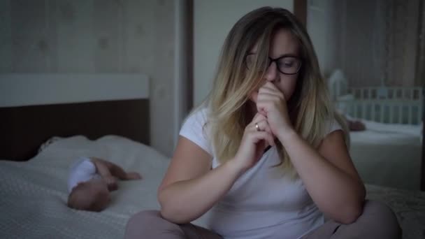 worried woman in eyeglasses suffers from headachesits on background of baby on bed at home - Filmmaterial, Video