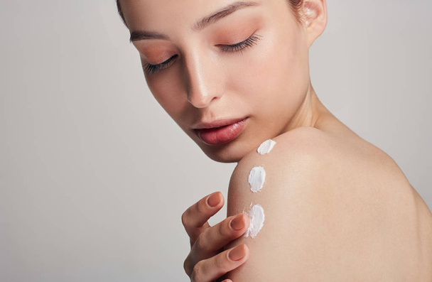 A young girl with clean fresh skin is applying cream on her shoulder with smears. Portrait on a light background. Beauty and care concept. - Valokuva, kuva