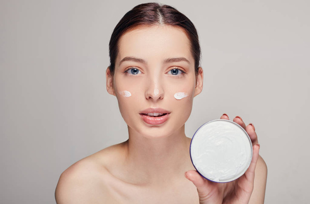 Young beautiful woman applying moisturizing cream on her face. Photo of young girl with flawless skin on background. - Foto, Imagem