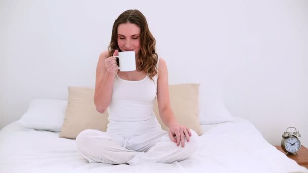 Pretty model sitting on bed drinking mug of coffee - Materiał filmowy, wideo