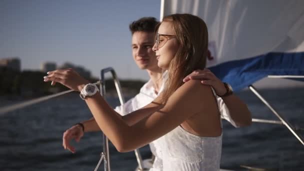 若いカップルのサイドビューはヨットで一緒に時間を過ごす。ボートの端に彼女のボーイフレンドと一緒に座って、水平線に一緒にポイント白いドレスとサングラスの女の子 - 映像、動画