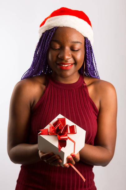 Beautiful Afro American girl in dress and Santa hat is holding p - 写真・画像