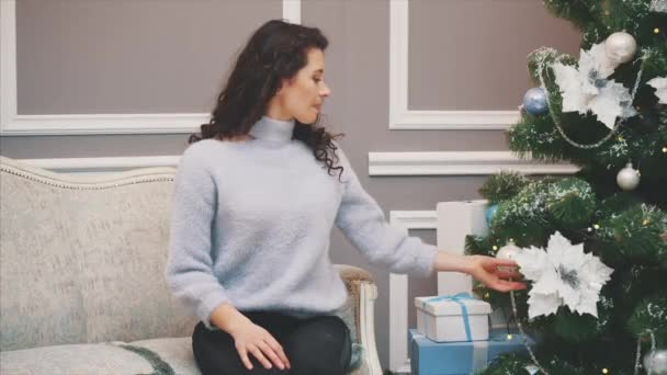 Pretty young woman is sitting on a sofa near Christmas tree, enjoying holiday spirit. - Filmmaterial, Video