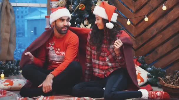 Cheerful women and men in red clothes, sitting on plaid carpet, covered with warm blanket, while lights are sparkling on the christmas tree. - Filmagem, Vídeo