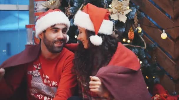 Happy man and woman sitting near Christmas tree, wrapped in red blanket, touching noses. - Filmati, video