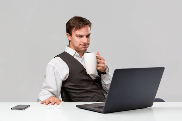 Office worker in a white t-shirt and grey vest is shocked checking information on laptop sitting at the white table drinking coffee. - Photo, Image