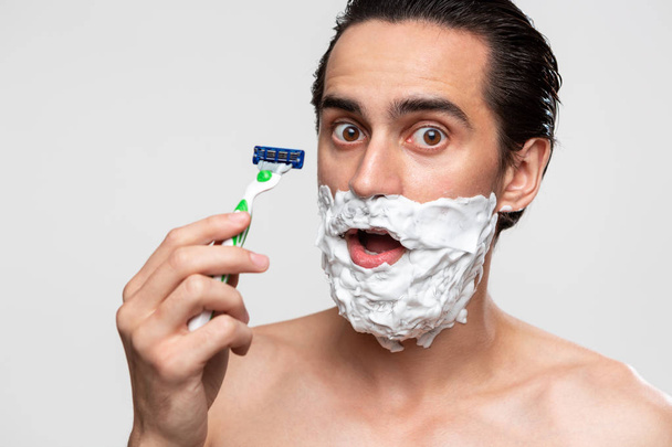 Bıyıklı yakışıklı adam sakalını kesmek için sopa ve tıraş köpüğü kullanıyor. Beyaz arka planda tek başına duruyor.. - Fotoğraf, Görsel