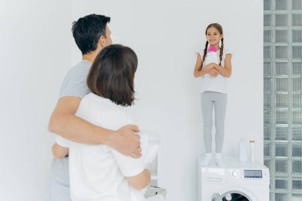 Back view of husband and wife embrace and talk to small girl standing on washer with bottle of detergent, do washing together during day off, have much work about house. Laundry day, home routine - Photo, Image