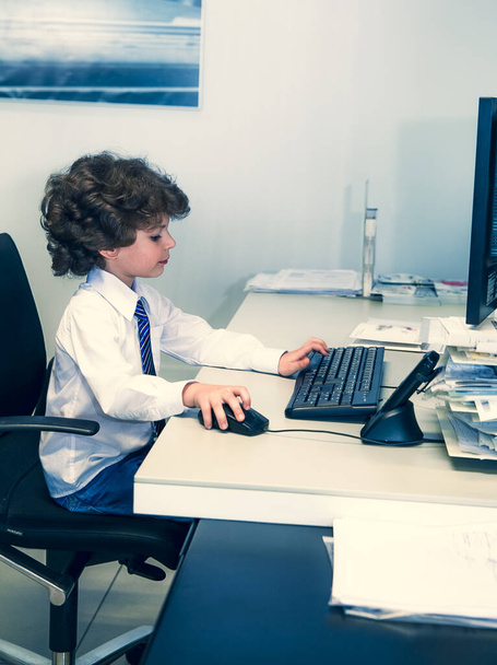Cute beautiful little boy children play as office worker sitting on working desk using business computer and working hard in a modern office - Zdjęcie, obraz