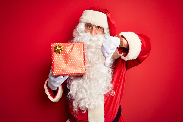 Hombre de mediana edad con traje de Papá Noel sosteniendo regalo sobre fondo rojo aislado con cara enojada, signo negativo que muestra aversión con pulgares hacia abajo, concepto de rechazo
 - Foto, Imagen