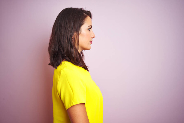 Młoda piękna kobieta w żółtym t-shirt stoi na różowym odizolowanym tle patrząc na bok, relaks profil pozować z naturalną twarz z pewnym uśmiechem. - Zdjęcie, obraz