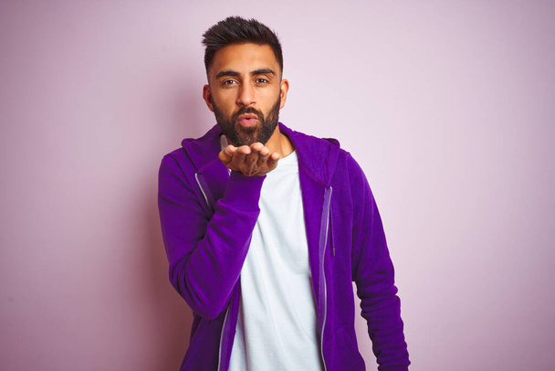 Young indian man wearing purple sweatshirt standing over isolated pink background looking at the camera blowing a kiss with hand on air being lovely and sexy. Love expression. - 写真・画像