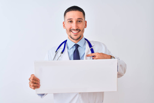 Young doctor man holding blank advertising banner over isolated background with surprise face pointing finger to himself - Foto, Bild