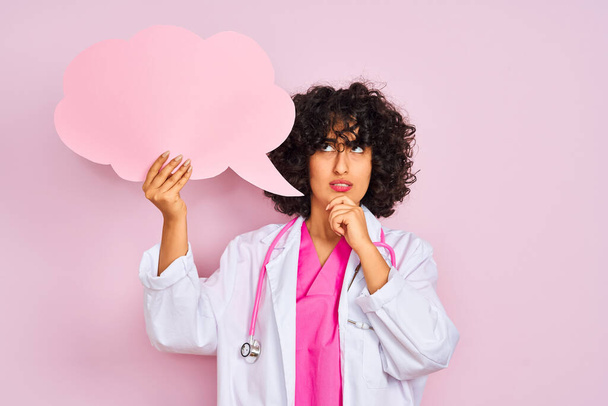 Mujer árabe joven con pelo rizado sosteniendo la burbuja del habla de la nube sobre el fondo rosa aislado cara seria pensando en la pregunta, idea muy confusa
 - Foto, imagen