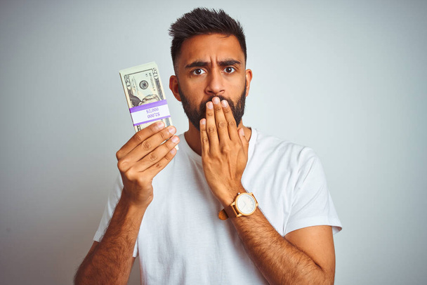 Young indian man holding dollars standing over isolated white background cover mouth with hand shocked with shame for mistake, expression of fear, scared in silence, secret concept - Фото, изображение