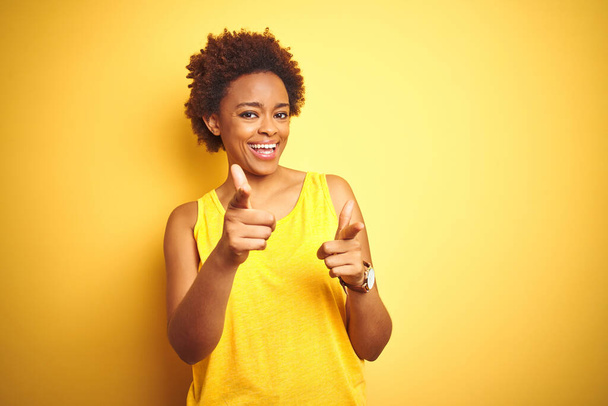 Beauitul africká americká žena v letním tričku přes izolované žluté pozadí ukazuje prsty na kameru s veselou a zábavnou tváří. Dobrá energie a vibrace. - Fotografie, Obrázek