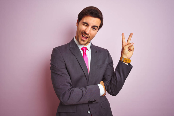 Молодой красивый бизнесмен в костюме и галстуке стоит на изолированном розовом фоне и улыбается счастливым лицом, подмигивая в камеру, делая знак победы. Номер два:
. - Фото, изображение