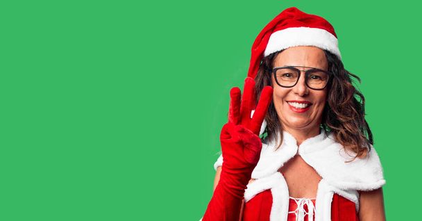 Женщина средних лет в костюме Санта-Клауса показывает и показывает пальцами номер три, улыбаясь уверенно и счастливо
. - Фото, изображение