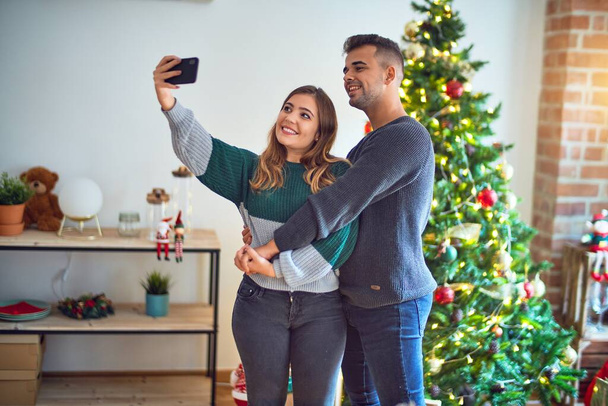 Young beautiful couple smiling happy and confident. Standing and hugging make selfie by camera around christmas tree at home - Foto, afbeelding