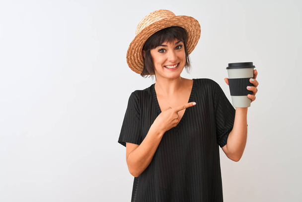 Hermosa mujer con sombrero de verano beber llevar café sobre fondo blanco aislado muy feliz señalando con la mano y el dedo
 - Foto, imagen