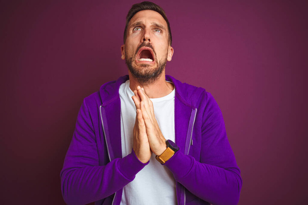 Young fitness man wearing casual sports sweatshirt over purple isolated background begging and praying with hands together with hope expression on face very emotional and worried. Asking for forgiveness. Religion concept. - Photo, Image