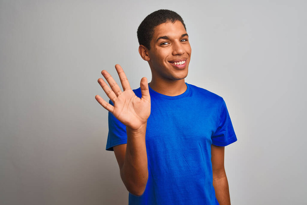 Mladý hezký arabský muž v modrém tričku stojící nad izolovaným bílým pozadím ukazující a ukazující nahoru s prsty číslo pět a usmívající se sebevědomě a šťastně. - Fotografie, Obrázek