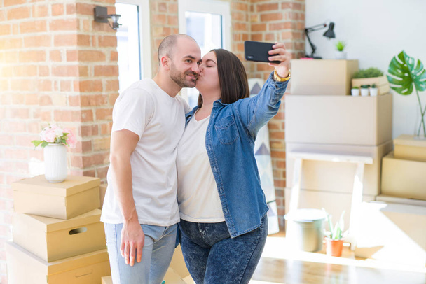 新居でスマホを使って写真を撮る若いカップル、新しいアパートに引っ越して嬉しそうに笑顔 - 写真・画像