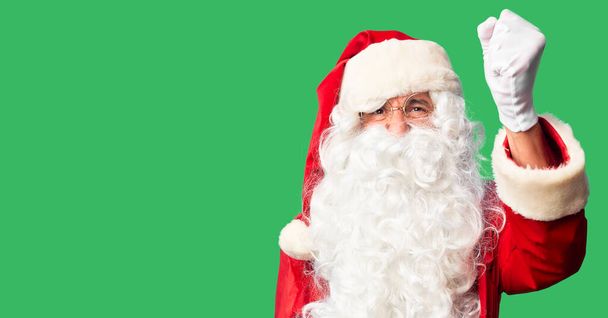 Красивый мужчина средних лет в костюме Санта-Клауса и с бородкой стоит злой и безумный, поднимая кулак, расстроенный и разъяренный, крича от злости. Ярость и агрессивность
. - Фото, изображение