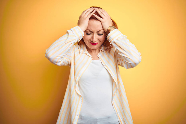 Belle femme rousse portant une chemise rayée debout sur fond jaune isolé souffrant de maux de tête désespérés et stressés parce que la douleur et la migraine. Mains sur la tête
. - Photo, image