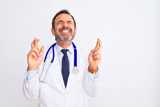 Middle age doctor man wearing coat and stethoscope standing over isolated white background gesturing finger crossed smiling with hope and eyes closed. Luck and superstitious concept. - Fotografie, Obrázek