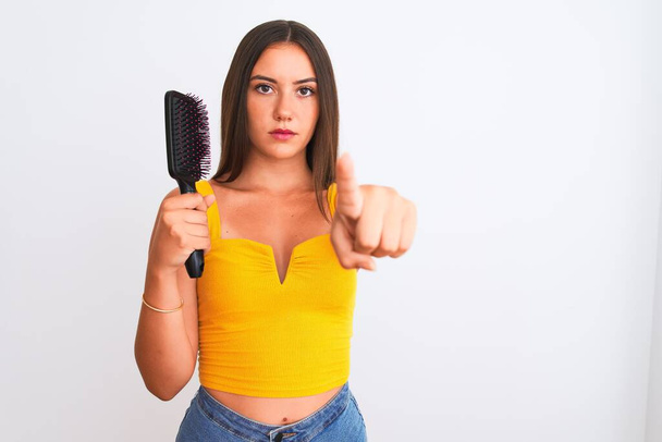 Young beautiful girl holding hair comb standing over isolated white background pointing with finger to the camera and to you, hand sign, positive and confident gesture from the front - 写真・画像