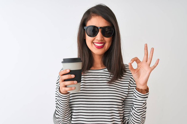 Chińska kobieta w okularach przeciwsłonecznych zabrać kawę na odizolowane białe tło robi ok znak palcami, doskonały symbol - Zdjęcie, obraz