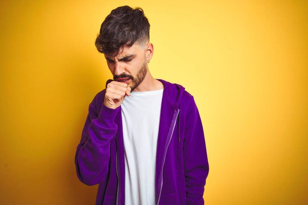 Giovane uomo con tatuaggio che indossa felpa viola sport su sfondo giallo isolato sensazione di malessere e tosse come sintomo per il freddo o bronchite. Concetto sanitario
. - Foto, immagini