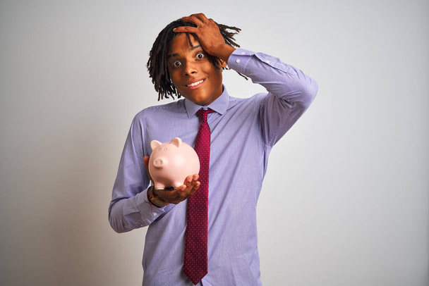 Afro americký obchodník s dredy drží prasátko přes izolované bílé pozadí vystresované s rukou na hlavě, šokovaný hanbou a překvapenou tváří, rozzlobený a frustrovaný. Strach a zmatek z omylu. - Fotografie, Obrázek