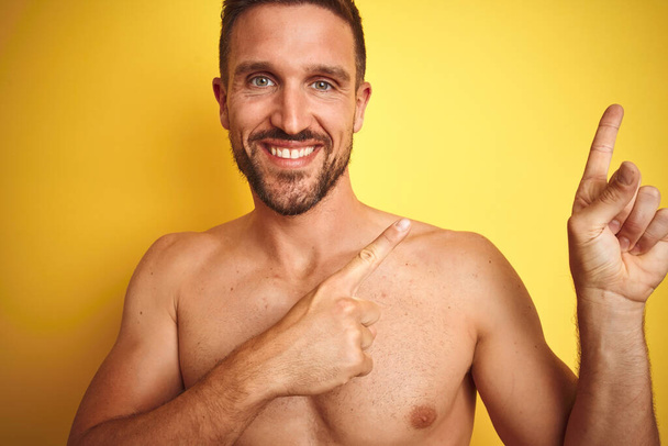 Сексуальний молодий без сорочки чоловік показує оголені фітнес-груди над жовтим ізольованим тлом, посміхаючись і дивлячись на камеру, спрямовану двома руками і пальцями в сторону
. - Фото, зображення