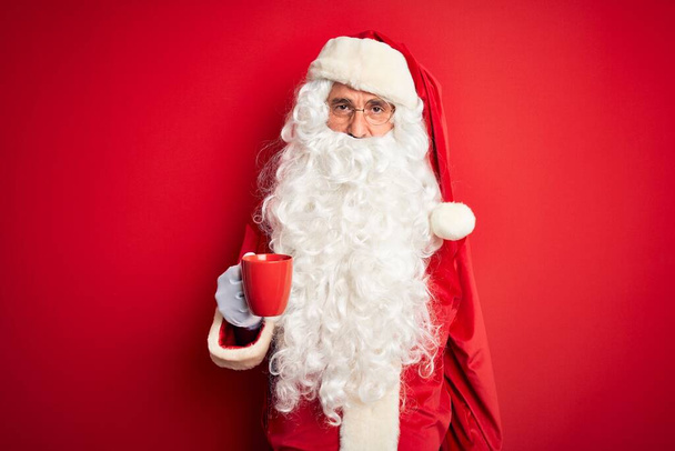 Старшеклассник в костюме Санта-Клауса держит чашку кофе на изолированном красном фоне с уверенным выражением на умном лице, думая серьезно
 - Фото, изображение