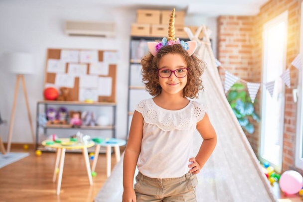 Hermoso niño usando gafas y diadema de unicornio de pie y sonriendo en el jardín de infantes
 - Foto, imagen