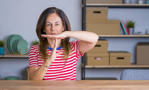 Старша жінка середнього віку сидить за столом вдома Робить час жестом руками, розчарованим і серйозним обличчям
 - Фото, зображення