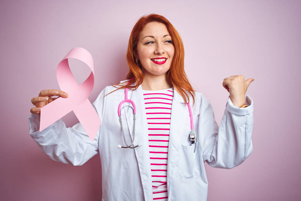 若いです赤毛医師女性保持癌リボン上のピンク隔離された背景ポインティングと表示上の親指で側に幸せな顔笑顔 - 写真・画像