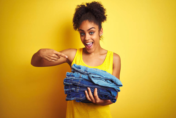 Young african american woman holding stack of jeans over isolated yellow background very happy pointing with hand and finger - Foto, imagen