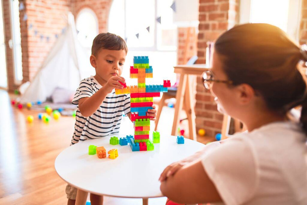 Beautiful teacher and toddler boy playing with construction blocks bulding tower at kindergarten - Fotó, kép