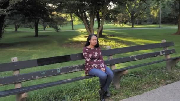 Yavaş çekim 180 derece yürür güzel bir Afrikalı Amerikalı kadının etrafında yürür eskimiş siyah bir ahşap ve beton bir park bankında oturur ağaçlar, binalar, bahçeler ve ötesinde güneş ışığı vardır.. - Video, Çekim