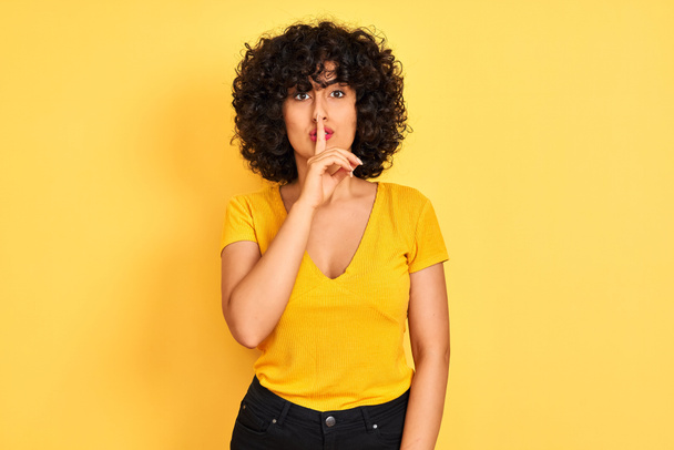 Jeune femme arabe aux cheveux bouclés portant un t-shirt debout sur un fond jaune isolé demandant de se taire avec le doigt sur les lèvres. Silence et concept secret
. - Photo, image