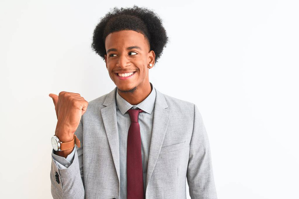 Młody afrykański biznesmen w garniturze stojący nad odizolowanym białym tłem uśmiechający się ze szczęśliwym wyglądem twarzy i wskazujący na bok z kciukiem w górze. - Zdjęcie, obraz