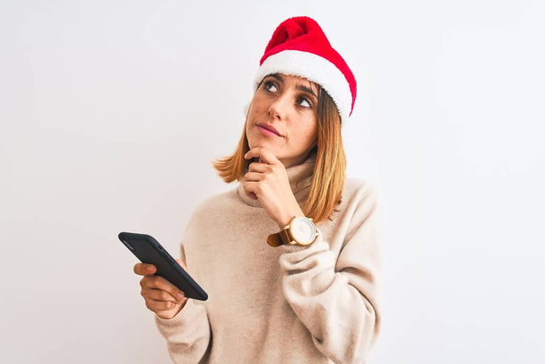 Beautiful redhead woman wearing christmas hat and using smartphone serious face thinking about question, very confused idea - Фото, изображение