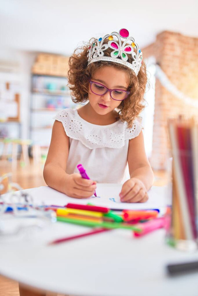 Piękny maluch nosi okulary i księżniczka korona siedzi rysunek przy użyciu papieru i markera pióra w przedszkolu - Zdjęcie, obraz