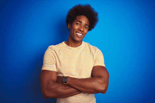 American man with afro hair wearing striped yellow t-shirt over isolated blue background happy face smiling with crossed arms looking at the camera. Positive person. - Photo, Image