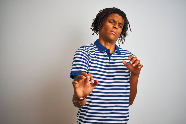 Afro man with dreadlocks wearing striped blue polo standing over isolated white background disgusted expression, displeased and fearful doing disgust face because aversion reaction. With hands raised. Annoying concept. - Фото, изображение